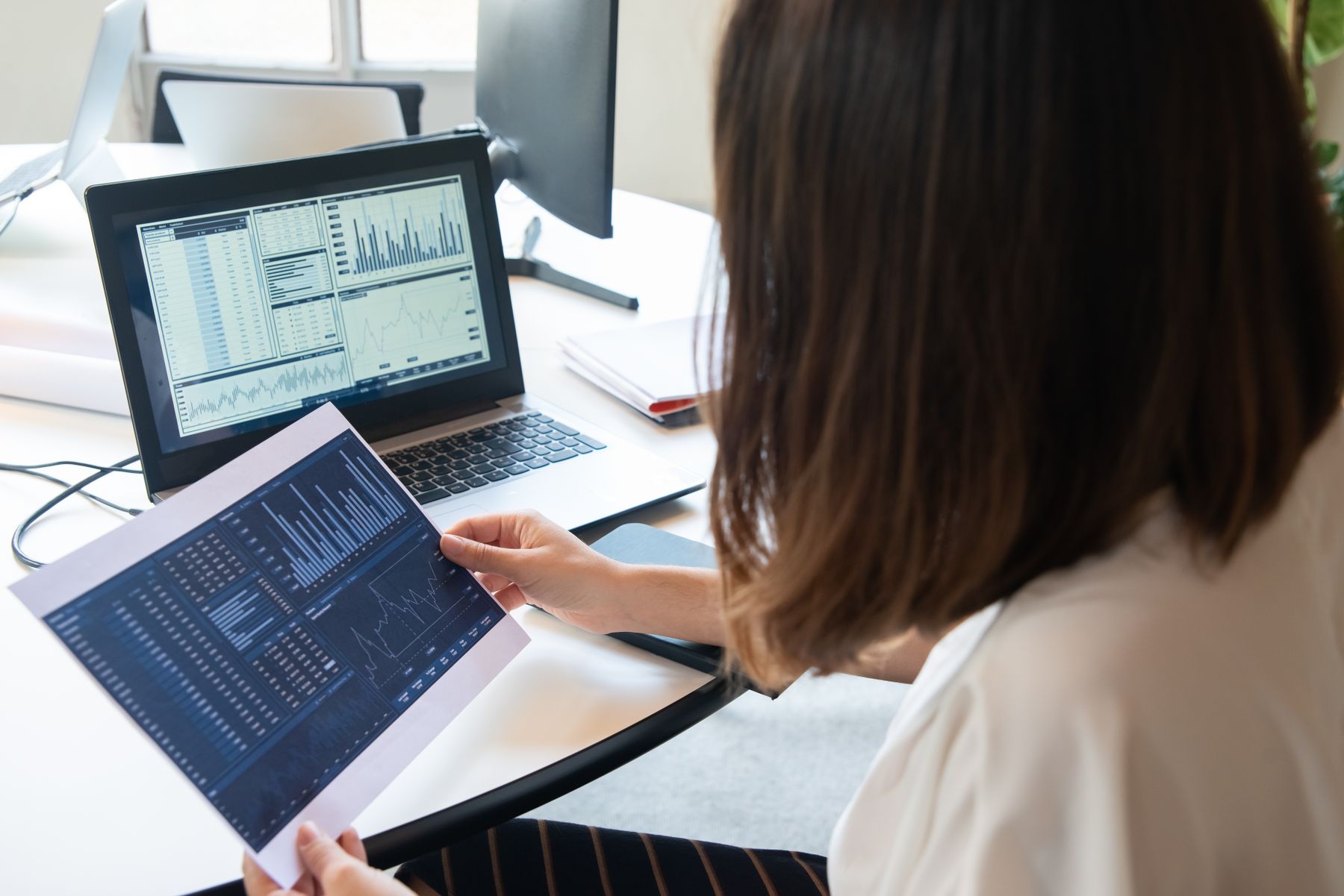Person comparing a printed chart report with tables and charts on a laptop.