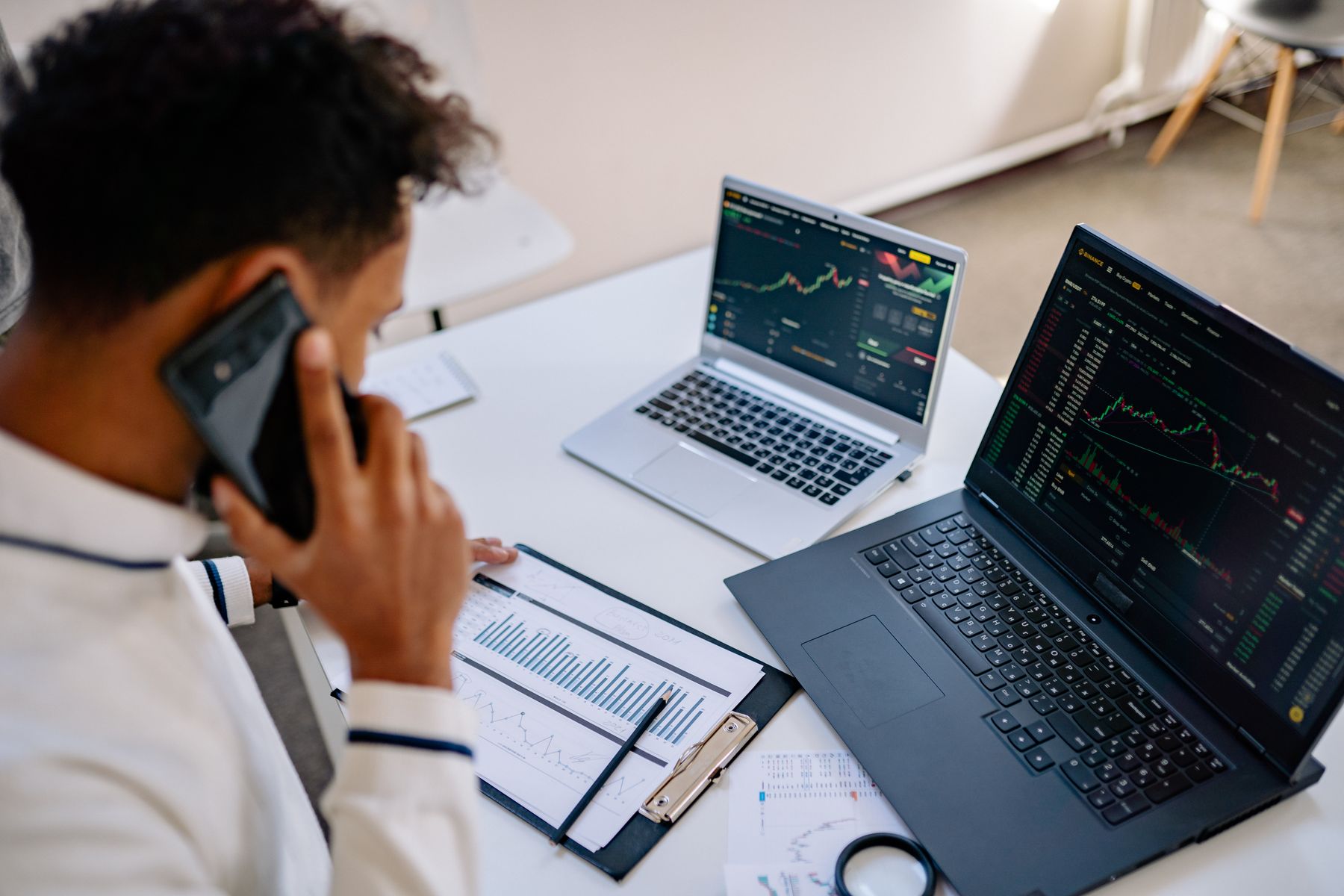 Person on the phone at a desk with two market-chart laptops and printed graphs.