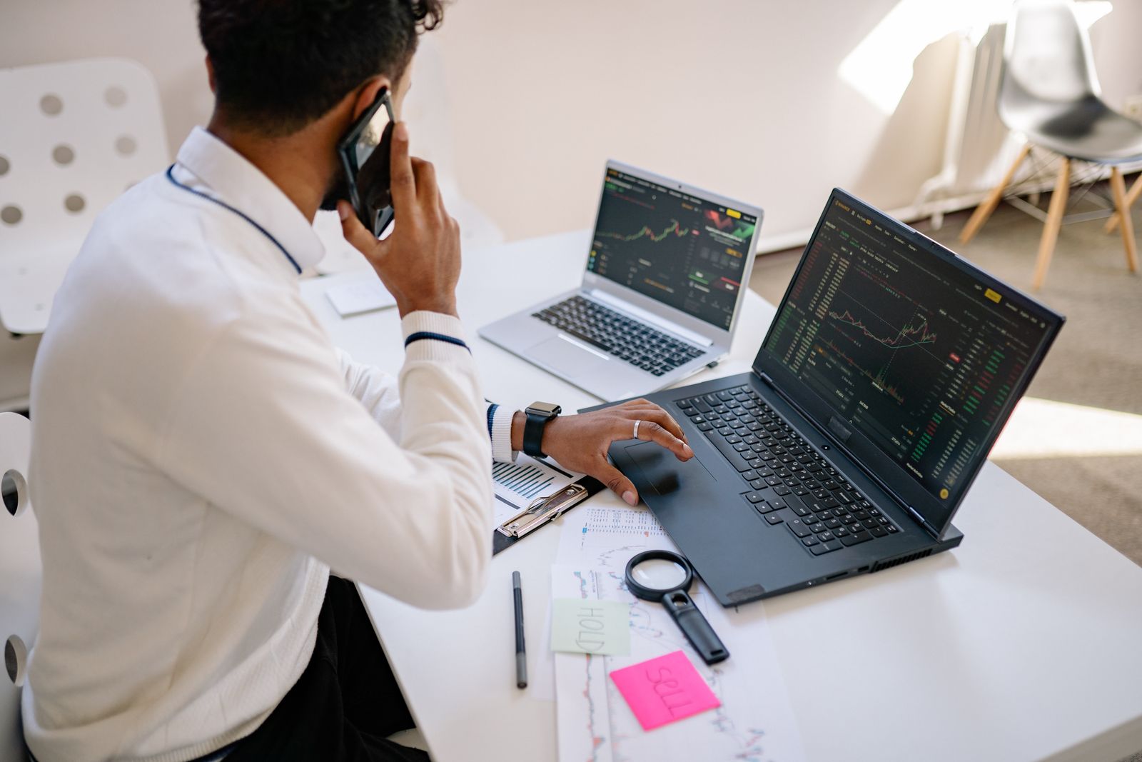 Person on the phone at a desk with two market-chart laptops and paper notes.